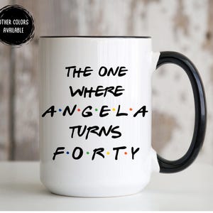 Op de afbeelding: Witte keramische mok met een zwarte handgreep en rand. De mok heeft de tekst "THE ONE WHERE ANGELA TURNS FORTY" in het zwart, met het woord "ANGELA" in kleurrijke stippen. Een zwarte cirkel met "OTHER COLORS AVAILABLE" staat linksboven.
