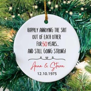 May include: White round Christmas ornament with black text that reads "Happily annoying the shit out of each other for 50 years... and still going strong!" with the names "Anna & Steven" and the date "12.10.1975" in red.