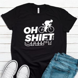 May include: Black t-shirt with the white text "OH SHIFT" and a bicycle graphic. The shirt is paired with blue jeans and white sneakers, set against a white wooden background. A casual, sporty theme.