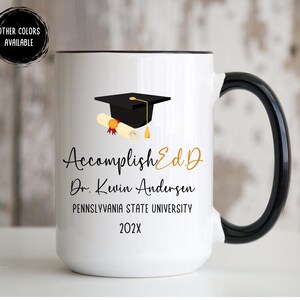 May include: White ceramic mug with a black handle and rim. The mug features a graduation cap and diploma graphic, along with the text "AccomplishEd.D Dr. Kevin Andersen Pennsylvania State University 202X". The mug has a black handle and rim.