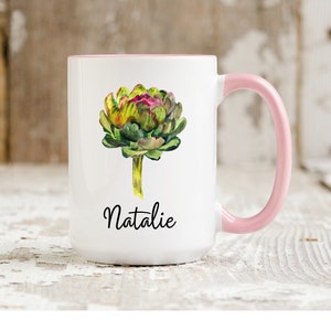 May include: White ceramic mug with a pink handle. The mug features a watercolor illustration of an artichoke and the name "Natalie" printed in black.