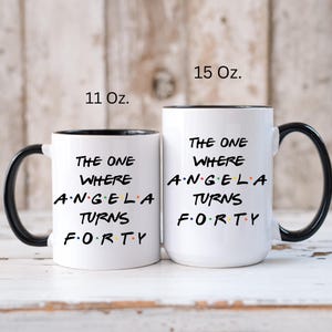 Op de afbeelding: Twee witte keramische mokken met zwarte handvatten en randen. De mokken hebben de tekst "THE ONE WHERE ANGELA TURNS FORTY" in zwart, met het woord "FORTY" in kleurrijke letters. E&eacute;n mok is 312 ml, de andere 425 ml.