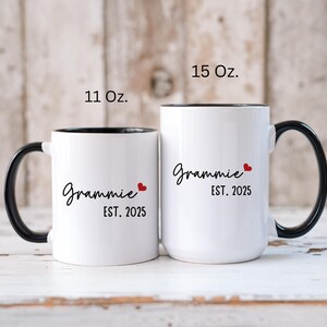 May include: Two white ceramic coffee mugs with black handles and black rims. The mugs have the text "Grammy Est. 2025" with a red heart. The mug on the left is 11 oz and the mug on the right is 15 oz.