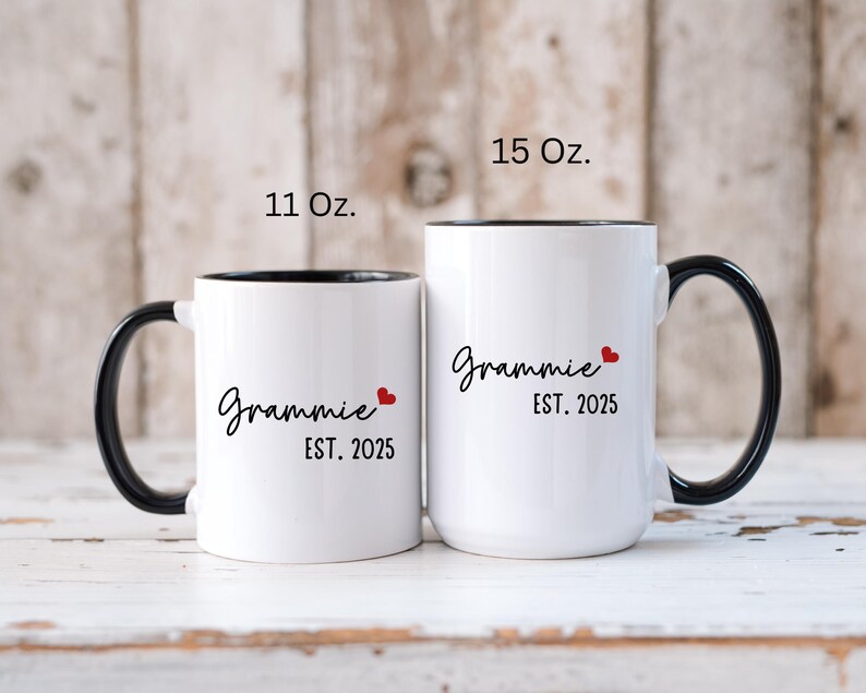 May include: Two white ceramic coffee mugs with black handles and black rims. The mugs have the text "Grammy Est. 2025" with a red heart. The mug on the left is 11 oz and the mug on the right is 15 oz.