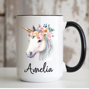 May include: White ceramic mug with a black handle. The mug features a watercolor illustration of a unicorn with a floral crown and the name "Amelia" printed below.