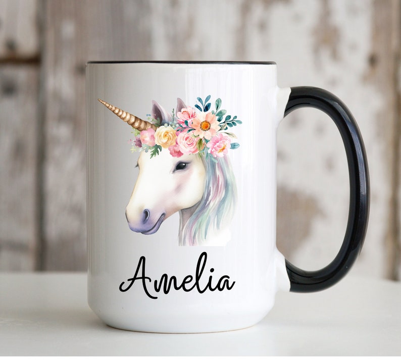 May include: White ceramic mug with a black handle. The mug features a watercolor illustration of a unicorn with a floral crown and the name "Amelia" printed below.