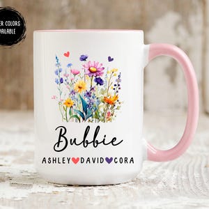 May include: White ceramic mug with a pink handle. The mug features a floral design with a variety of colorful flowers and the word "Bubbie" in black lettering. The text "Ashley David Cora" is printed below the word "Bubbie" with a heart between each name.
