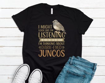 May include: Black t-shirt with the text "I MIGHT LOOK LIKE I'M LISTENING TO YOU BUT IN MY HEAD I'M THINKING ABOUT DARK-EYED JUNCOS" and a bird illustration. The shirt is paired with blue jeans and white sneakers.