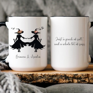 May include: White ceramic mug with a black handle and rim. The mug features a black silhouette of two witches holding hands, with the names "Bianca & Amelia" below. The other side has the text "Just a pinch of salt... and a whole lot of sass."
