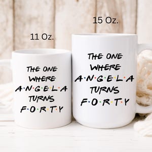 Op de afbeelding: Twee witte keramische mokken met de tekst "THE ONE WHERE ANGELA TURNS FORTY". De mokken zijn gemarkeerd met 11 Oz. en 15 Oz. De tekst is in het zwart, met het woord "ANGELA" in kleur.