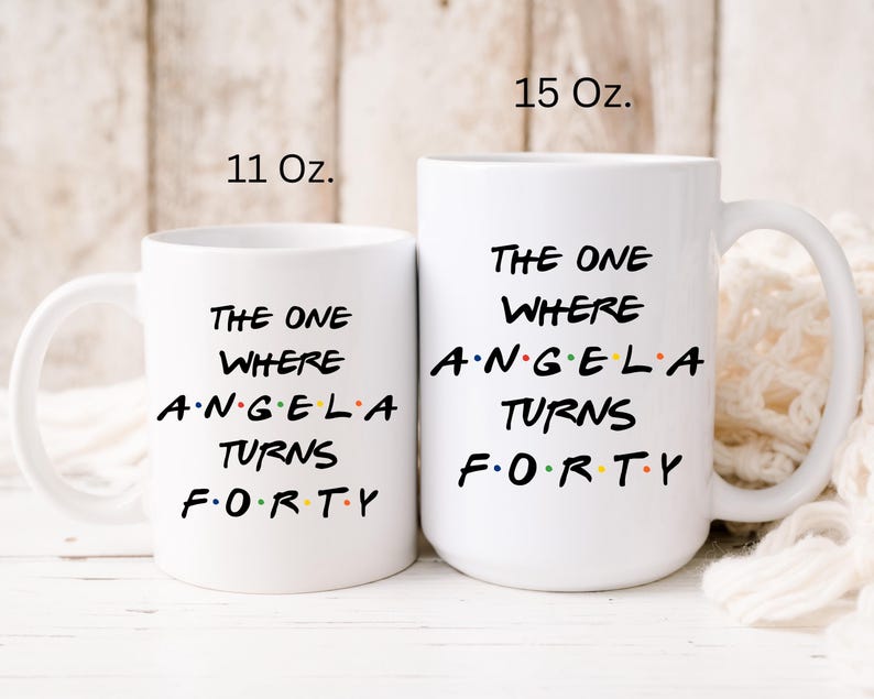 Op de afbeelding: Twee witte keramische mokken met de tekst "THE ONE WHERE ANGELA TURNS FORTY". De mokken zijn gemarkeerd met 11 Oz. en 15 Oz. De tekst is in het zwart, met het woord "ANGELA" in kleur.