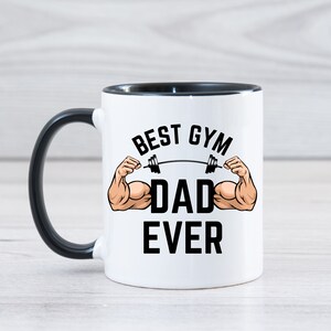 May include: White ceramic coffee mug with a black handle and black rim. The mug has a graphic of a muscular arm lifting a barbell with the text "Best Gym Dad Ever".