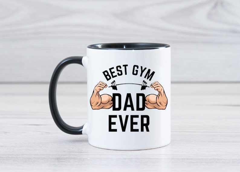 May include: White ceramic coffee mug with a black handle and black rim. The mug has a graphic of a muscular arm lifting a barbell with the text "Best Gym Dad Ever".