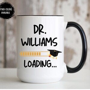 May include: White coffee mug with a black handle. The mug has black text that reads "Dr. Williams Loading..." with a graduation cap graphic above the text.
