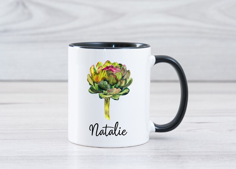 May include: White ceramic mug with a black rim and handle. The mug features a watercolor illustration of an artichoke with the name "Natalie" printed below.