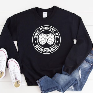 May include: Black sweatshirt with the text "THE PURSUIT OF HOPPINESS" in a white circular design. The design includes two white hop cones. White sneakers and blue jeans are also in the image.
