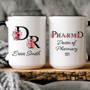 May include: Two white ceramic mugs with black handles and black rims. The mug on the left has a floral design and the letters "DR" with the name "Erica Smith" below. The mug on the right has the text "PHARM.D. Doctor of Pharmacy 202X".