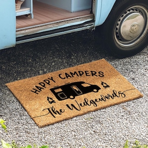 May include: A rectangular coir doormat with the words "HAPPY CAMPERS" and "The Wedgewoods" in black script. A black camper illustration is centered on the mat, with small campfire icons on either side. The mat is placed in front of a blue camper van.
