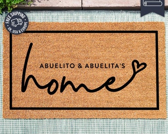 Puede incluir: Una alfombra de coco marrón con un borde negro y el texto "ABUELITO & ABUELITA'S home" en una fuente cursiva con un corazón al final. La alfombra está sobre una superficie de azulejos grises y blancos.