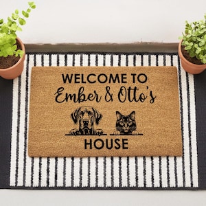 Peut inclure: Un paillasson rectangulaire en fibre de coco avec le texte "WELCOME TO Ember & Otto's HOUSE". Le tapis présente des illustrations d'un chien et d'un chat. Le tapis est sur un tapis rayé avec des plantes en pot de chaque côté.