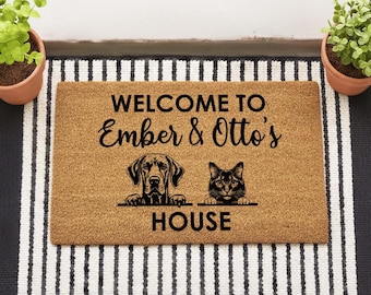 May include: A rectangular coir doormat with the text "WELCOME TO Ember & Otto's HOUSE". The mat features illustrations of a dog and a cat. The mat is on a striped rug with potted plants on either side.