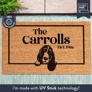 May include: A brown coir doormat with a black border and the text "The Carrolls EST. 1986". A black and white illustration of a basset hound dog peeking over the top of the mat.