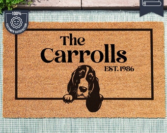 May include: A brown coir doormat with a black border and the text "The Carrolls EST. 1986". A black and white illustration of a basset hound dog peeking over the top of the mat.