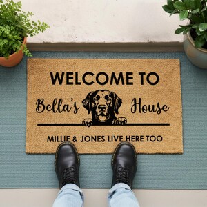K&ouml;nnte beinhalten: Eine rechteckige Fu&szlig;matte mit dem Text "WELCOME TO Bella's House" und einer Hunde-Grafik. Unter der Grafik steht "MILLIE & JONES LIVE HERE TOO". Die Matte ist beige mit schwarzem Text und einer schwarzen Hunde-Grafik.