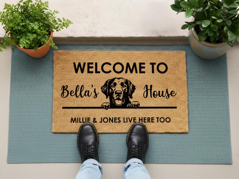 K&ouml;nnte beinhalten: Eine rechteckige Fu&szlig;matte mit dem Text "WELCOME TO Bella's House" und einer Hunde-Grafik. Unter der Grafik steht "MILLIE & JONES LIVE HERE TOO". Die Matte ist beige mit schwarzem Text und einer schwarzen Hunde-Grafik.