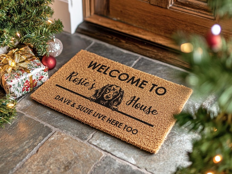 K&ouml;nnte beinhalten: Eine rechteckige Fu&szlig;matte in Braun mit dem Text "WELCOME TO Rosie's House" und "DAVE & SUZIE LIVE HERE TOO" in Schwarz, mit einer Dackel-Illustration. Die Matte liegt auf einer Steinoberfl&auml;che, mit Weihnachtsdekorationen im Hintergrund.