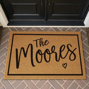 Puede incluir: Una alfombra de puerta rectangular hecha de fibra de coco natural. La alfombra presenta las palabras "The Moores" en una fuente negra y cursiva, con un peque&ntilde;o s&iacute;mbolo de coraz&oacute;n. Un borde negro enmarca el texto y la alfombra.