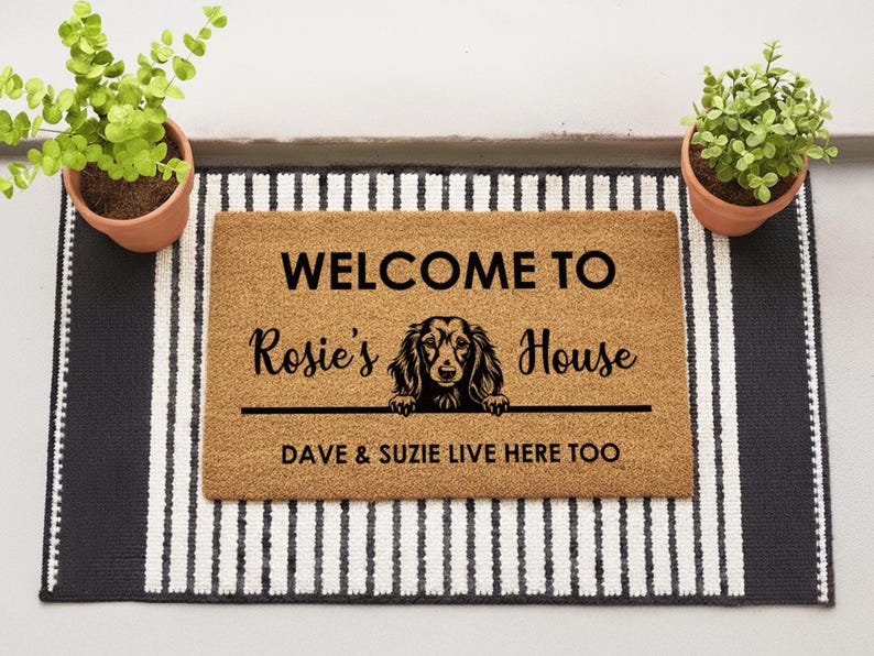K&ouml;nnte beinhalten: Eine rechteckige Fu&szlig;matte mit dem Text "WELCOME TO Rosie's House" und einer Dackel-Illustration. Die Matte liegt auf einem schwarz-wei&szlig; gestreiften Teppich, mit Topfpflanzen auf beiden Seiten. Der Text "DAVE & SUZIE LIVE HERE TOO" ist ebenfalls enthalten.