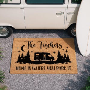 Pode incluir: Tapete retangular feito de fibra de coco natural. O tapete apresenta um gráfico preto de uma autocaravana, árvores, lua e estrelas. O texto diz "The Fischers" e "HOME IS WHERE YOU PARK IT".