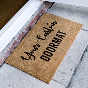 Puede incluir: Una alfombra de coco marr&oacute;n con el texto "Your Custom Doormat" en letras negras cursivas.