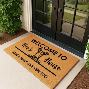 K&ouml;nnte beinhalten: Eine rechteckige Fu&szlig;matte mit beigem Hintergrund und schwarzem Text. Der Text lautet "WELCOME TO Neo's House" mit einer Hunde-Grafik und "JOSIE & MARK LIVE HERE TOO". Die Matte liegt vor einer schwarzen T&uuml;r.