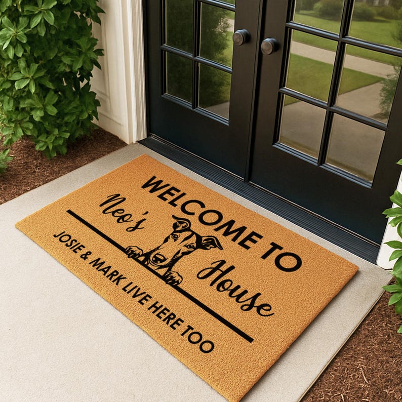 K&ouml;nnte beinhalten: Eine rechteckige Fu&szlig;matte mit beigem Hintergrund und schwarzem Text. Der Text lautet "WELCOME TO Neo's House" mit einer Hunde-Grafik und "JOSIE & MARK LIVE HERE TOO". Die Matte liegt vor einer schwarzen T&uuml;r.