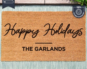 Puede incluir: Una alfombra de coco marrón con el texto "Happy Holidays" y "THE GARLANDS" en letras negras. La alfombra está hecha con tecnología UV Stick.