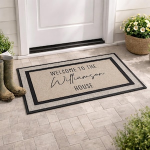 May include: A rectangular doormat with a tan centre and a black border. The text "WELCOME TO THE" is printed in a simple black font, followed by the name "Williamson" in a black cursive script, and the word "HOUSE" in a simple black font.
