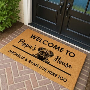 May include: A rectangular doormat with the text "WELCOME TO Pippa's House" and "MICHELLE & RYAN LIVE HERE TOO" in black. A pug illustration is centered on the mat. The mat is a natural tan color and sits on a brick surface.