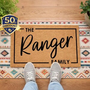 May include: A rectangular doormat with the text "THE Ranger FAMILY" in black script. The mat is on a patterned rug with a wooden floor background. A 50% discount badge is in the upper left corner.