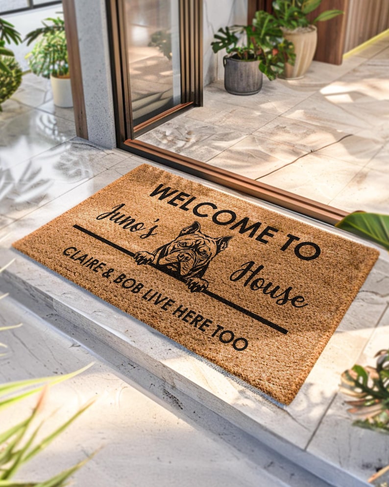 K&ouml;nnte beinhalten: Rechteckige Fu&szlig;matte aus Naturfasern mit dem Text "WELCOME TO House", "Juno's" und "CLAIRE & BOB LIVE HERE TOO". In der Mitte befindet sich eine schwarze Hundeillustration. Die Matte liegt vor einer T&uuml;r.