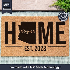May include: A brown coir doormat with the word "HOME" in black letters. The state of Arizona is outlined in black with the word "arizona" written in cursive inside the state. The text "EST. 2023" is written below the word "HOME".