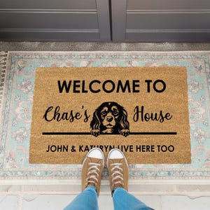 K&ouml;nnte beinhalten: Eine rechteckige Fu&szlig;matte mit dem Text "WELCOME TO Chase's House" und einer Hundeillustration. Darunter steht der Text "JOHN & KATHRYN LIVE HERE TOO". Die Matte liegt auf einem gemusterten Teppich, mit F&uuml;&szlig;en und Schuhen einer Person.