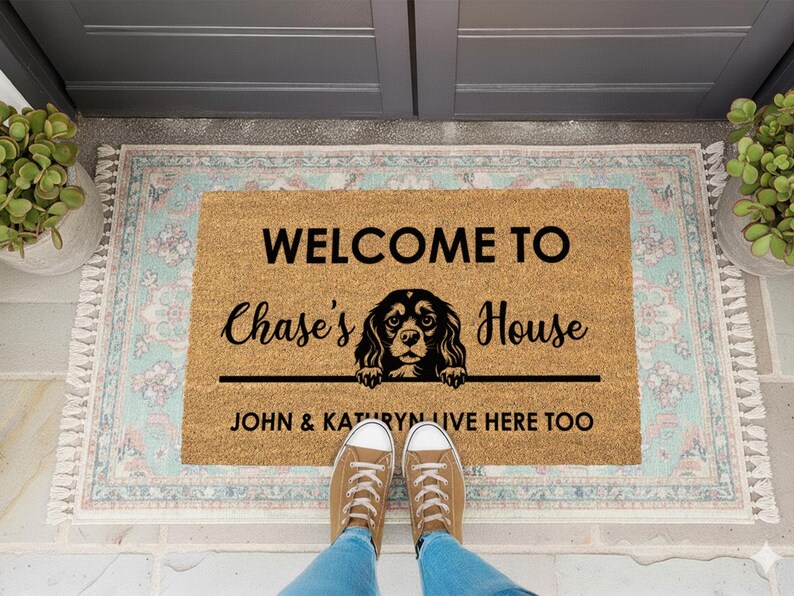 K&ouml;nnte beinhalten: Eine rechteckige Fu&szlig;matte mit dem Text "WELCOME TO Chase's House" und einer Hundeillustration. Darunter steht der Text "JOHN & KATHRYN LIVE HERE TOO". Die Matte liegt auf einem gemusterten Teppich, mit F&uuml;&szlig;en und Schuhen einer Person.