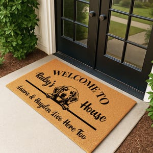 Op de afbeelding: Een rechthoekige deurmat met een bruine achtergrond en zwarte tekst. De tekst luidt "WELCOME TO House" met een hondenillustratie. Onder de hond staat "Ruby's" en "Laura & Hayden Live Here Too".