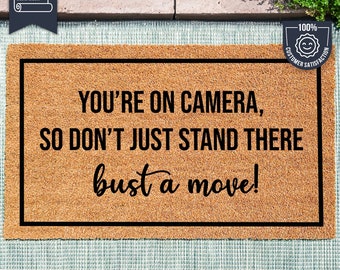 Może przedstawiać: Brązowa mata kokosowa z czarną obwódką i nadrukowanym tekstem "YOU'RE ON CAMERA, SO DON'T JUST STAND THERE bust a move!" Mata leży na jasnoniebieskiej powierzchni z białym wzorem.