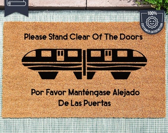 May include: A brown coir doormat with the text "Please Stand Clear Of The Doors" and a black silhouette of a train. The text "Por Favor Manténgase Alejado De Las Puertas" is also printed on the mat. The mat is made with UV Stick technology.