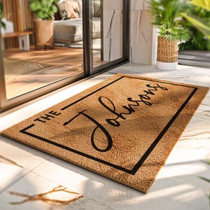 Puede incluir: Una alfombra rectangular hecha de fibras naturales. La alfombra tiene un borde negro y el apellido "The Johnsons" en una fuente elegante. La alfombra est&aacute; colocada sobre una superficie embaldosada cerca de una puerta de cristal.