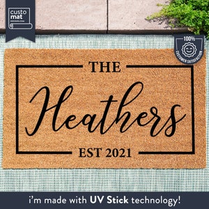 以下が含まれることがあります： 黒い文字で「THE Heathers EST 2021」と書かれた茶色のココナッツ繊維のドアマット。テキストは装飾的なフォントです。右上の隅には、小さな白黒のステッカーに「100％顧客満足」と書いてあります。
