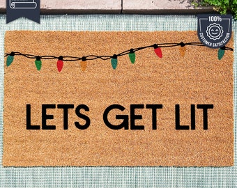 May include: A rectangular coir doormat with the phrase "LETS GET LIT" in black, and a string of colourful Christmas lights. The text "i'm made with UV Stick technology!" is at the bottom. The mat is made of natural coir fibre.
